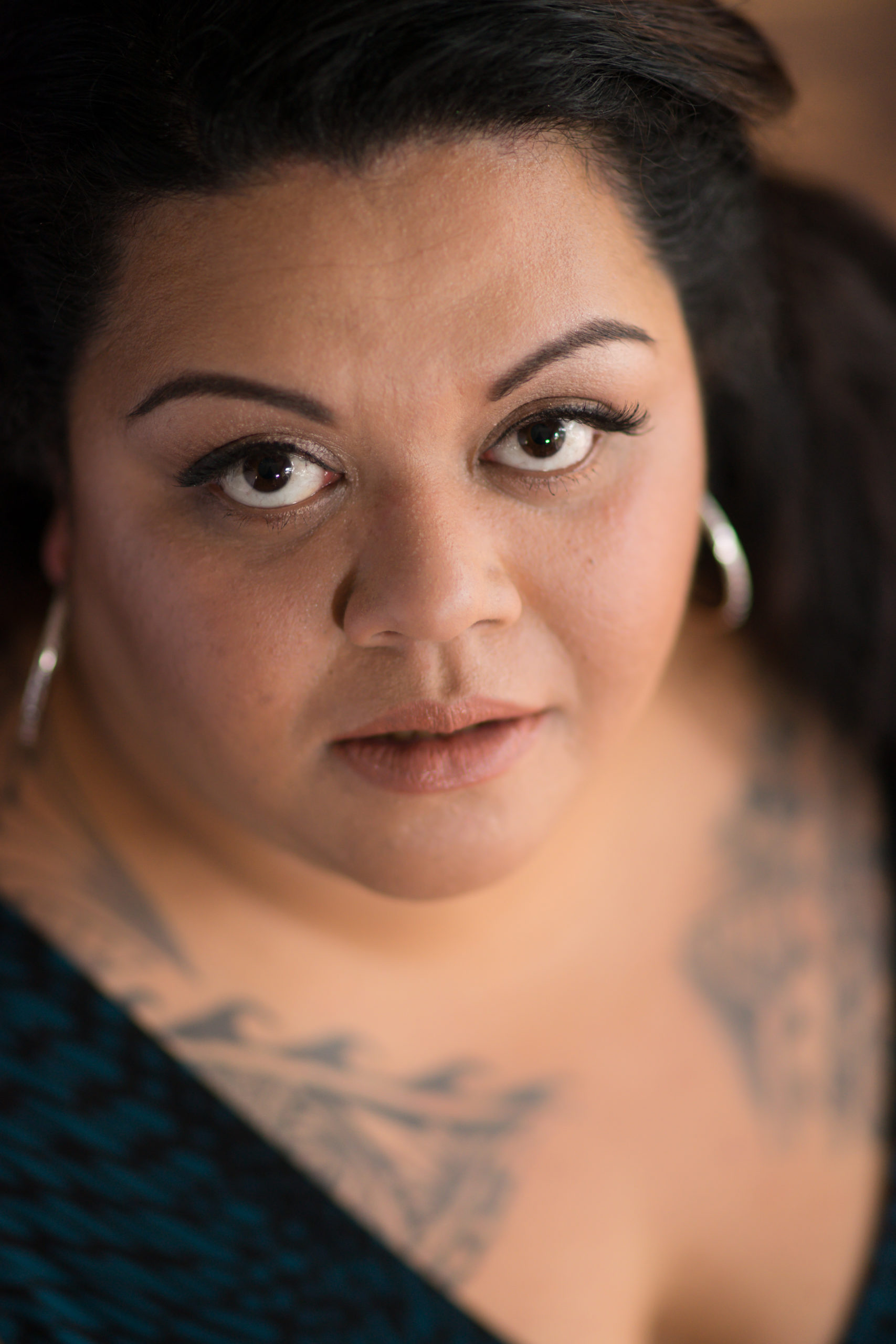 A fat Hawaiian woman gazes up at the camera in a close-up shot that shows her serious face, silver hoop earrings and traditional-style upper chest tattoos.