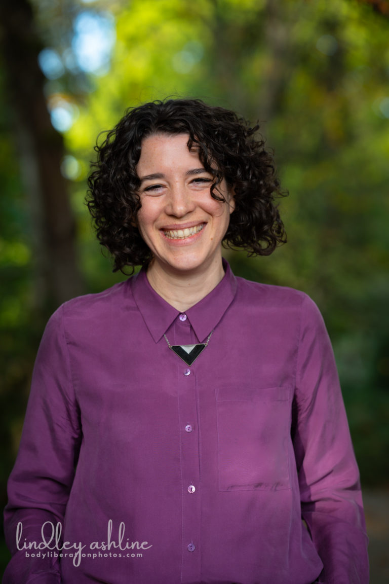 A thin pale-skinned woman with dark curly hair smiles widely in front of a background of trees and bits of sky. She's wearing a purple button-down shirt and triangle necklace